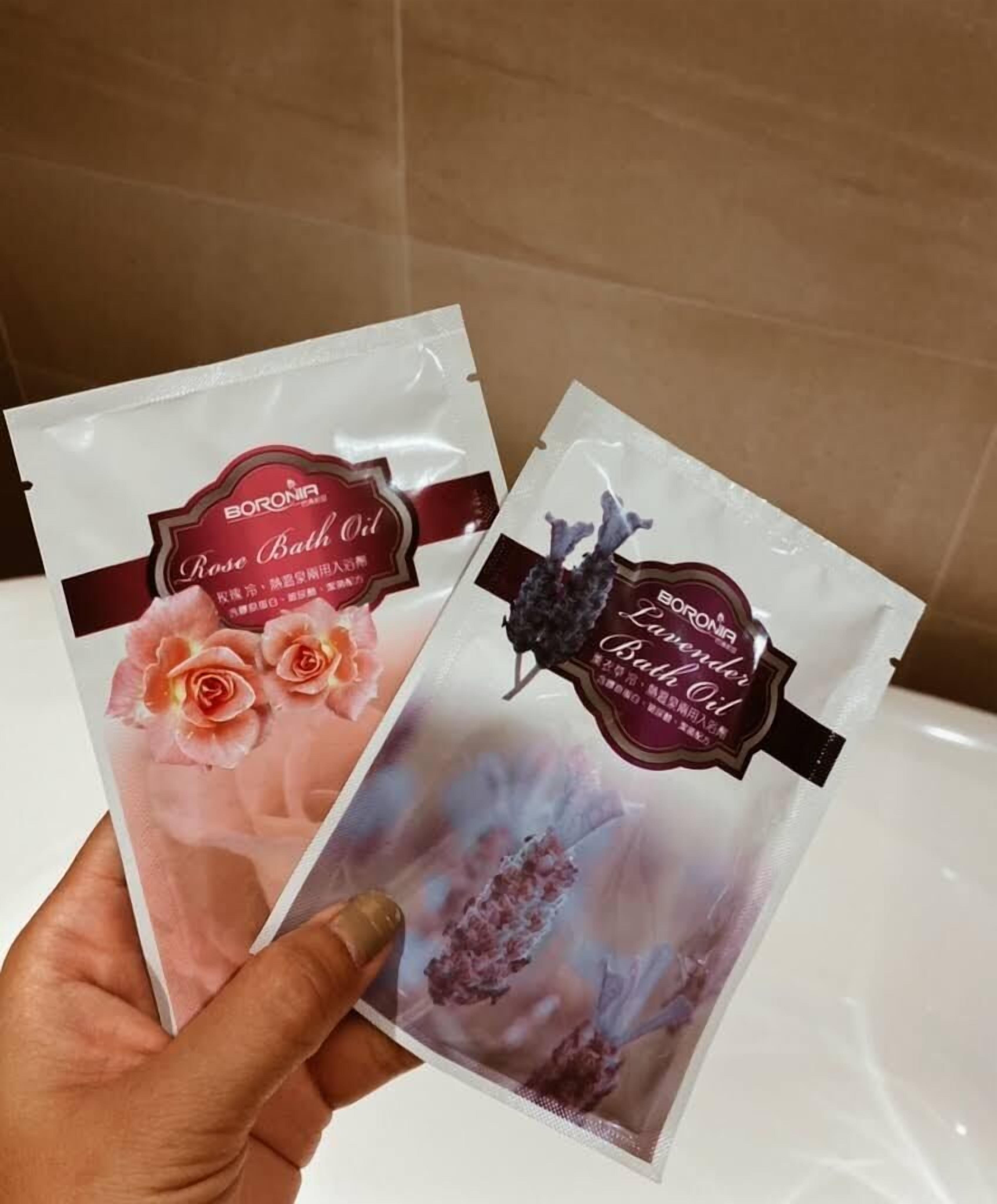 To pakker Boronia badeolie, hvoraf den ene er mærket 'Rose Badeolie' og den anden 'Lavendel Badeolie', holdt af en person.