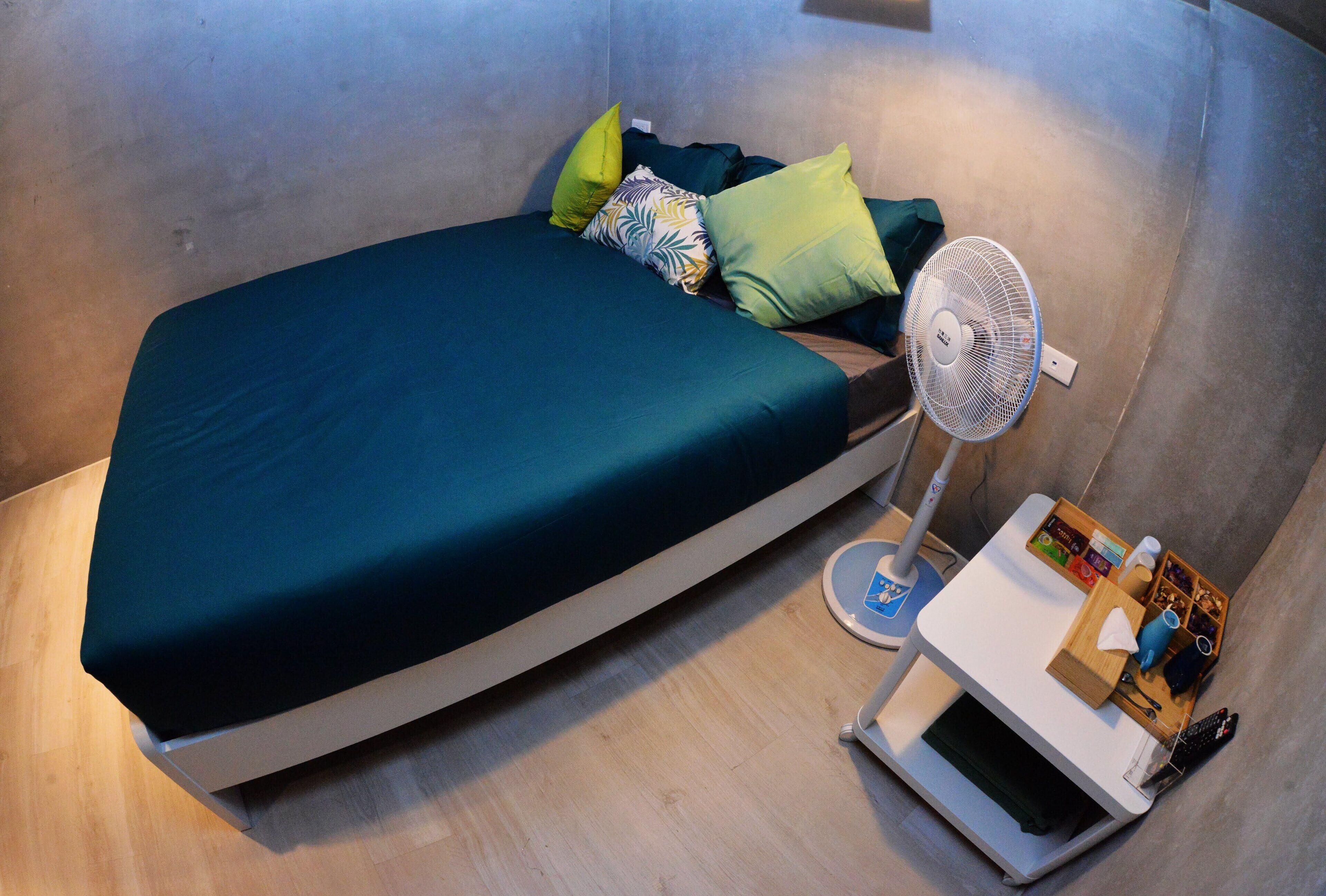 A small, modern room with a bed, a standing fan, and a small table with a remote control.