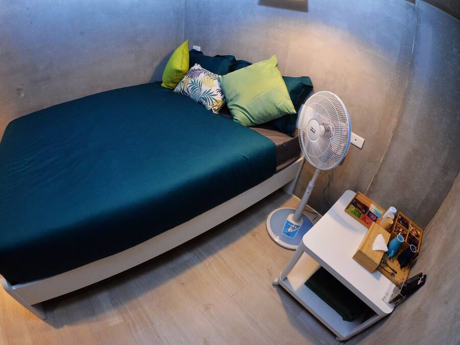 Une petite chambre moderne, équipée d’un lit, d’un ventilateur sur pied et d’une petite table sur laquelle se trouve une télécommande.