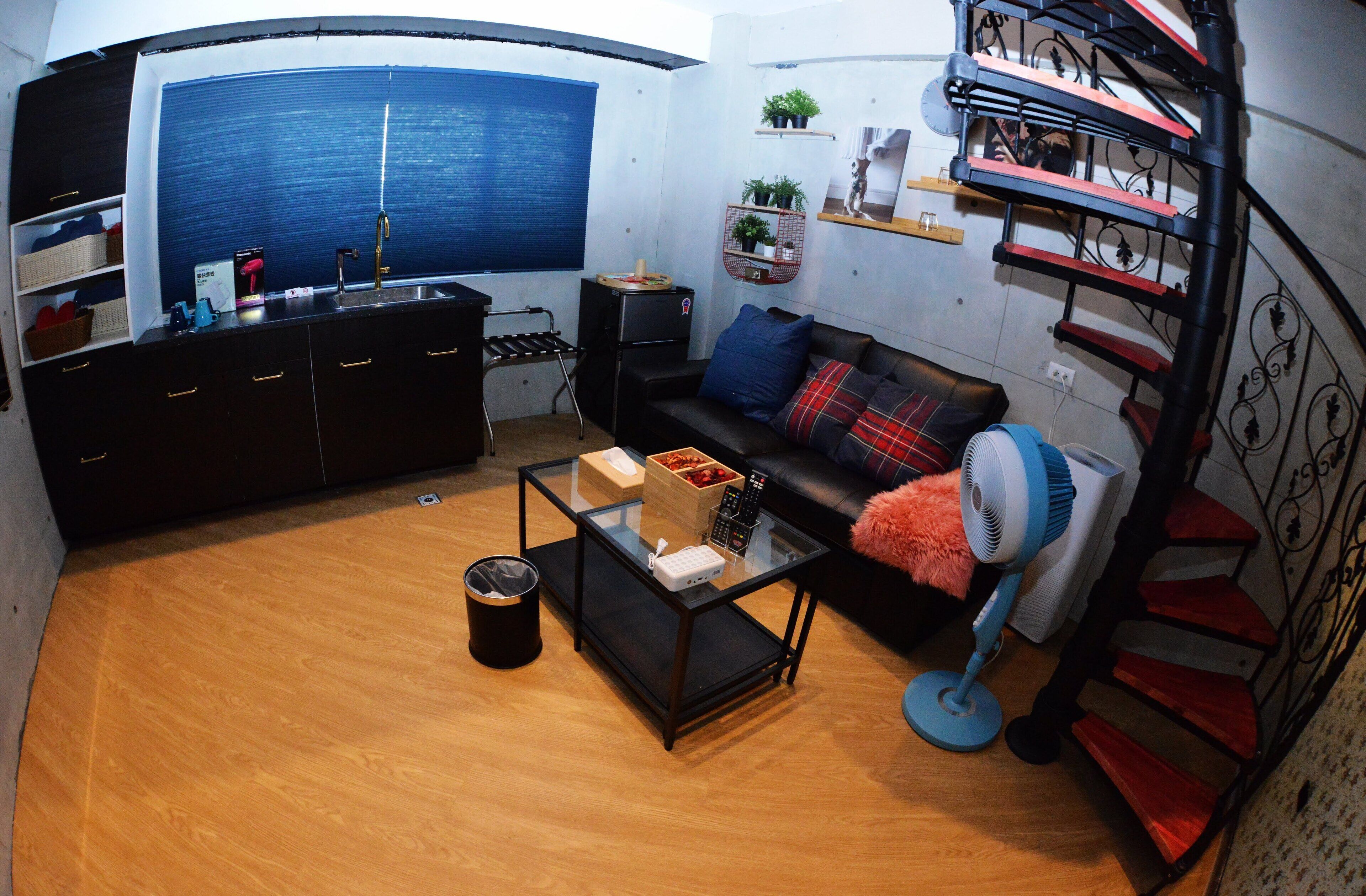A compact living space with a kitchenette, a sofa with cushions, a glass coffee table, a fan, and a spiral staircase.