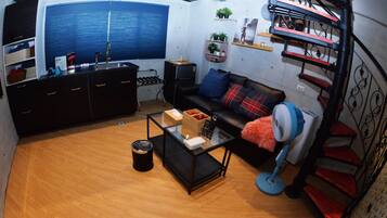 A compact living space with a kitchenette, a sofa with cushions, a glass coffee table, a fan, and a spiral staircase.