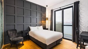 Ein modernes Hotelzimmer mit einem großen Bett, einem Sessel, einem Schreibtisch und Blick auf die Stadt durch ein großes Fenster.