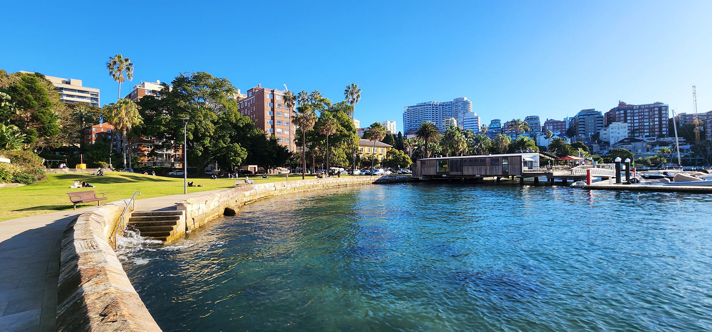 A waterfront park with a swimming pool, benches, and a wooden structure near the water.