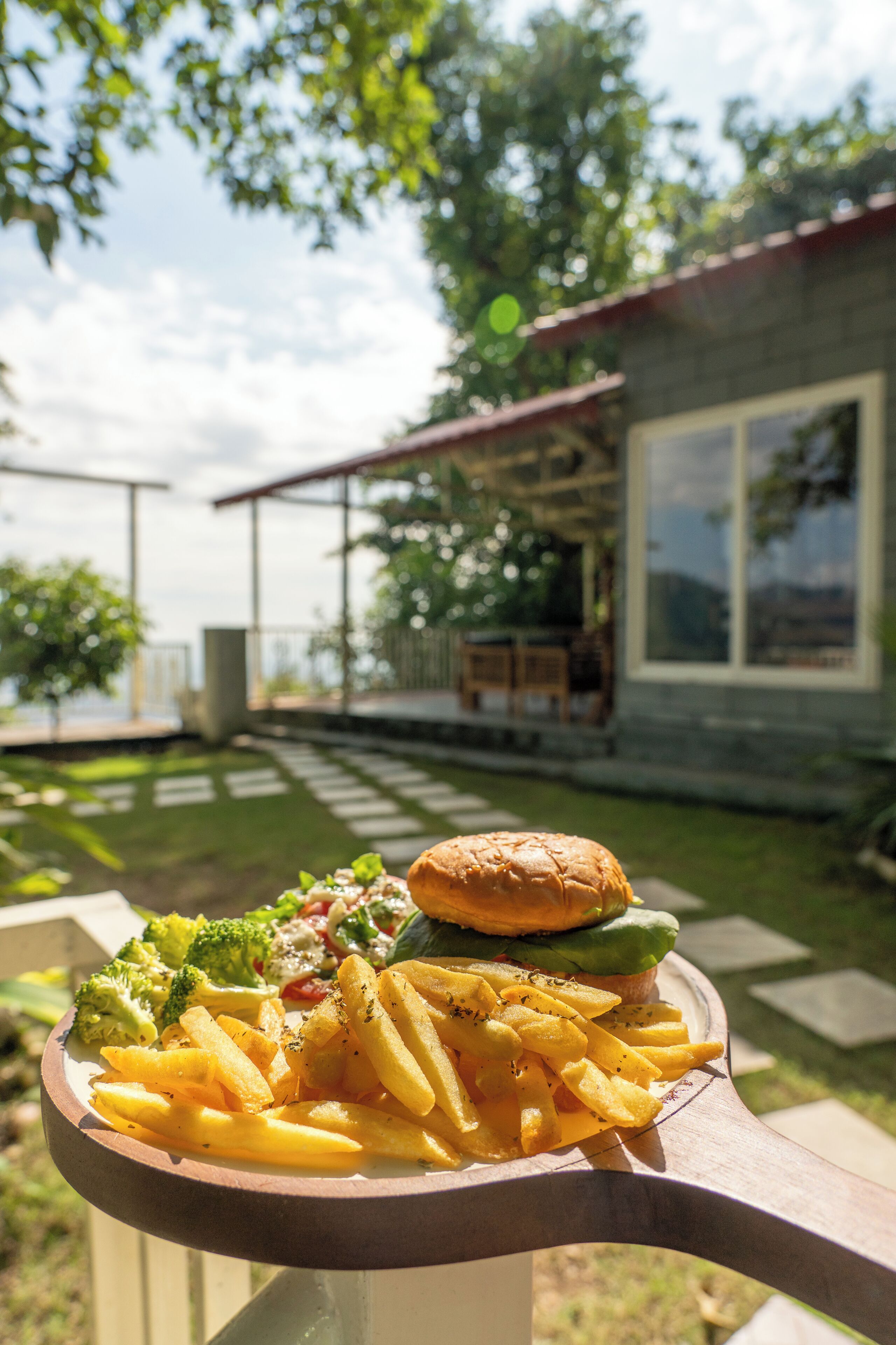 A meal of fries, burger, and vegetables on a wooden table with a view of a house and garden.