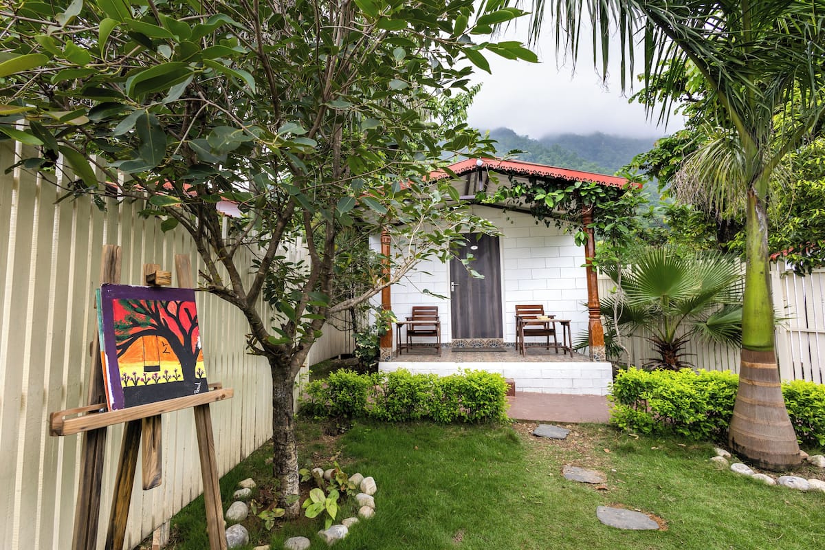 A small house with a porch, surrounded by a garden with a palm tree and a painting on an easel.