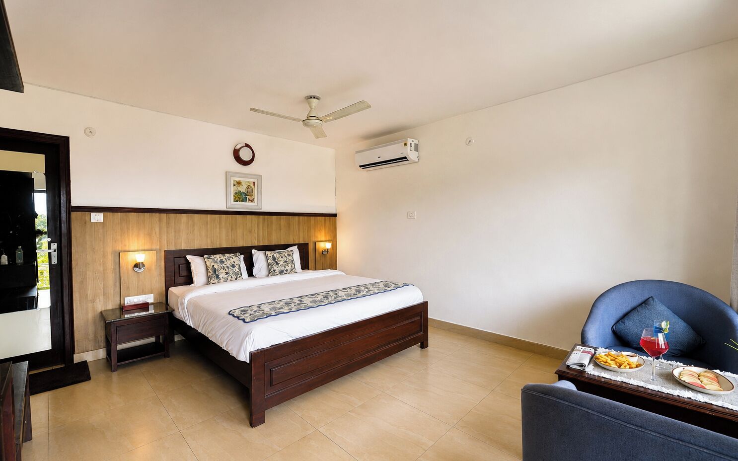A hotel room with a bed, bedside tables, a chair, a small table with food and drink, and a ceiling fan.