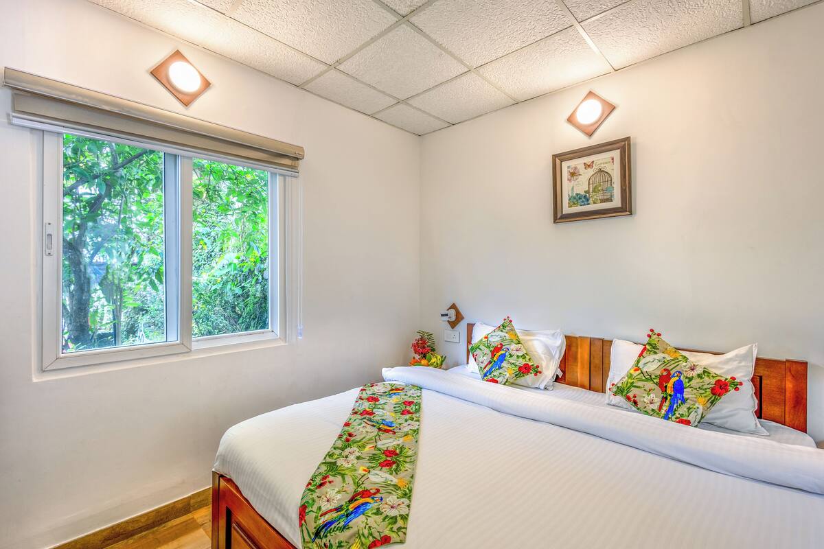 A neatly made bed with floral pillows and a matching blanket, a window with a view of greenery, and a framed picture on the wall.