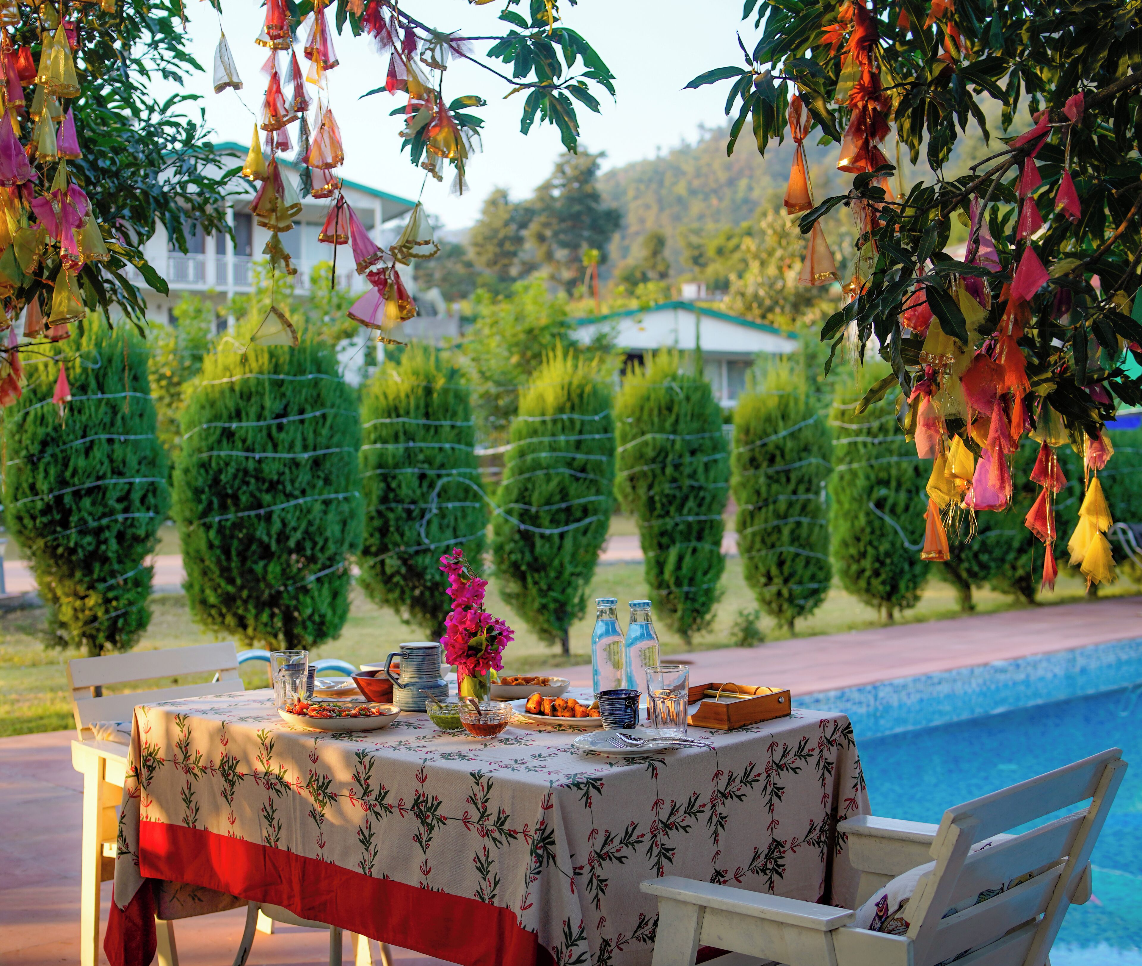 Mesa junto a la piscina con mantel floral, rodeada de setos bien cuidados y vista despejada a la alberca.