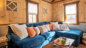 Ein gemütliches Zimmer mit einem blauen Ecksofa, Holzwänden und zwei Fenstern mit Jalousien.