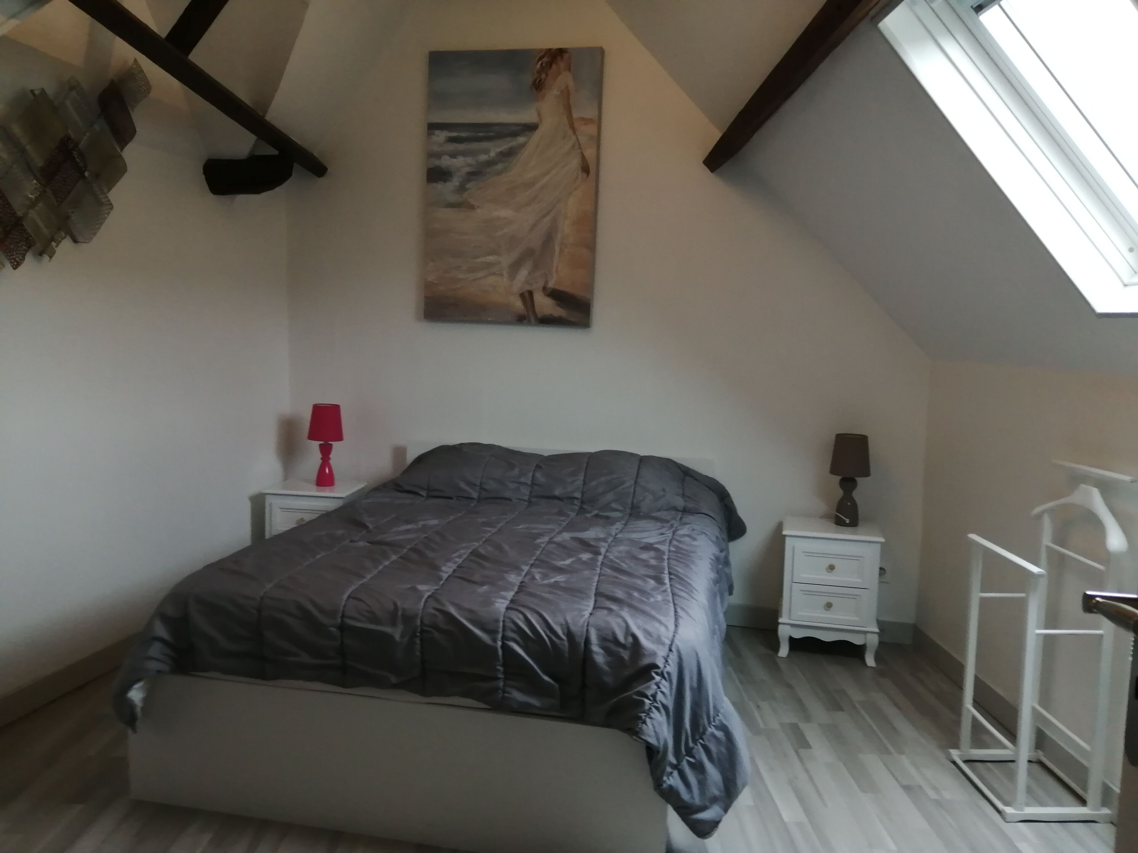 A bedroom with a bed, a nightstand, a lamp, a painting, and a skylight.