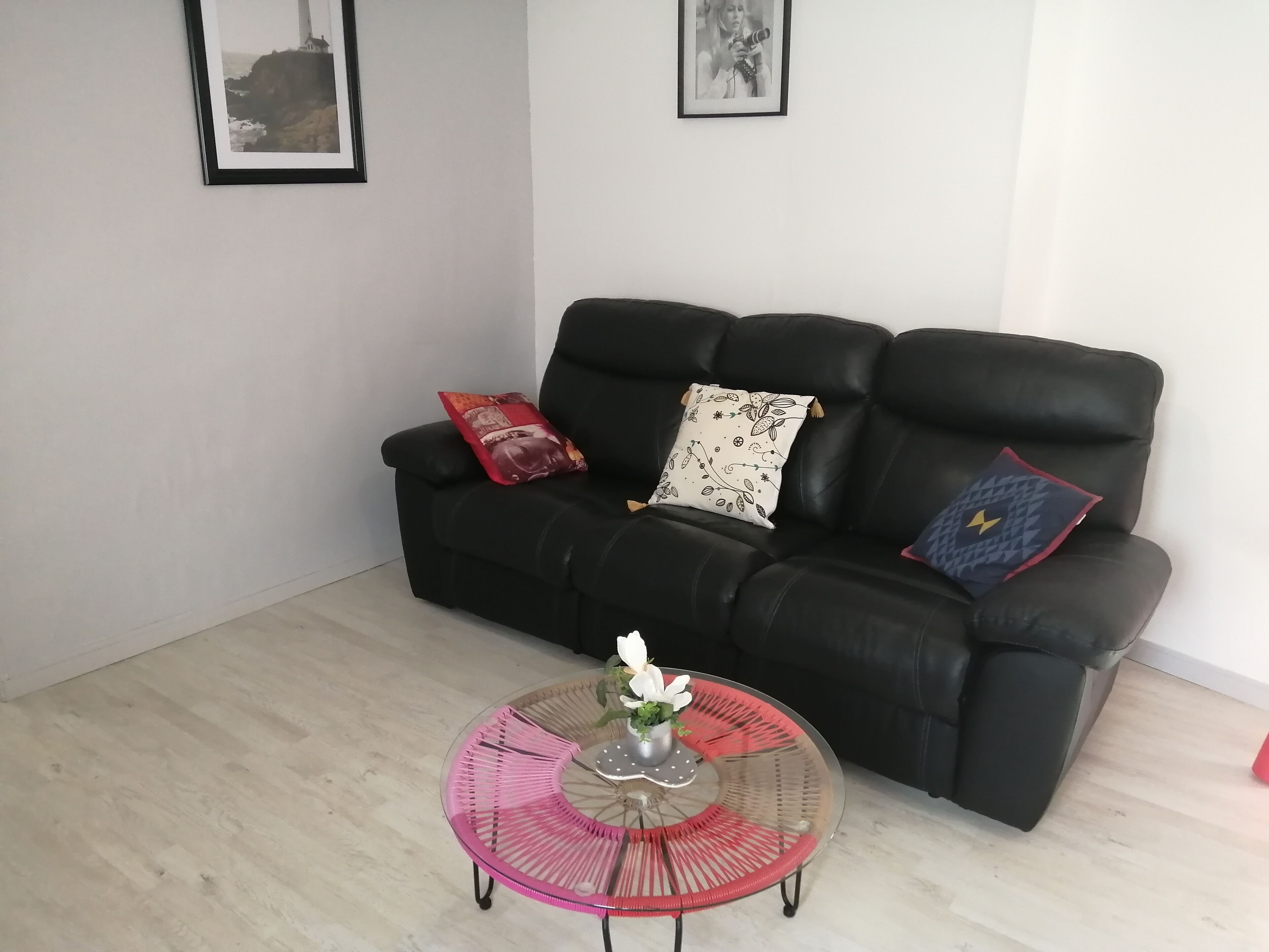 Een zwarte leren bank met drie gestreepte kussens, een ronde glazen salontafel met een witte bloemenvaas, en twee ingelijste foto’s aan de muur.