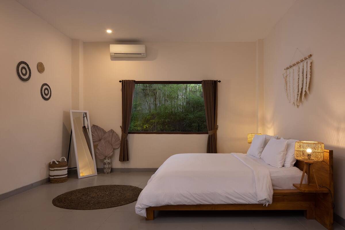 Um quarto com uma janela grande, uma cama com roupa de cama branca, um abajur e um espelho na parede.