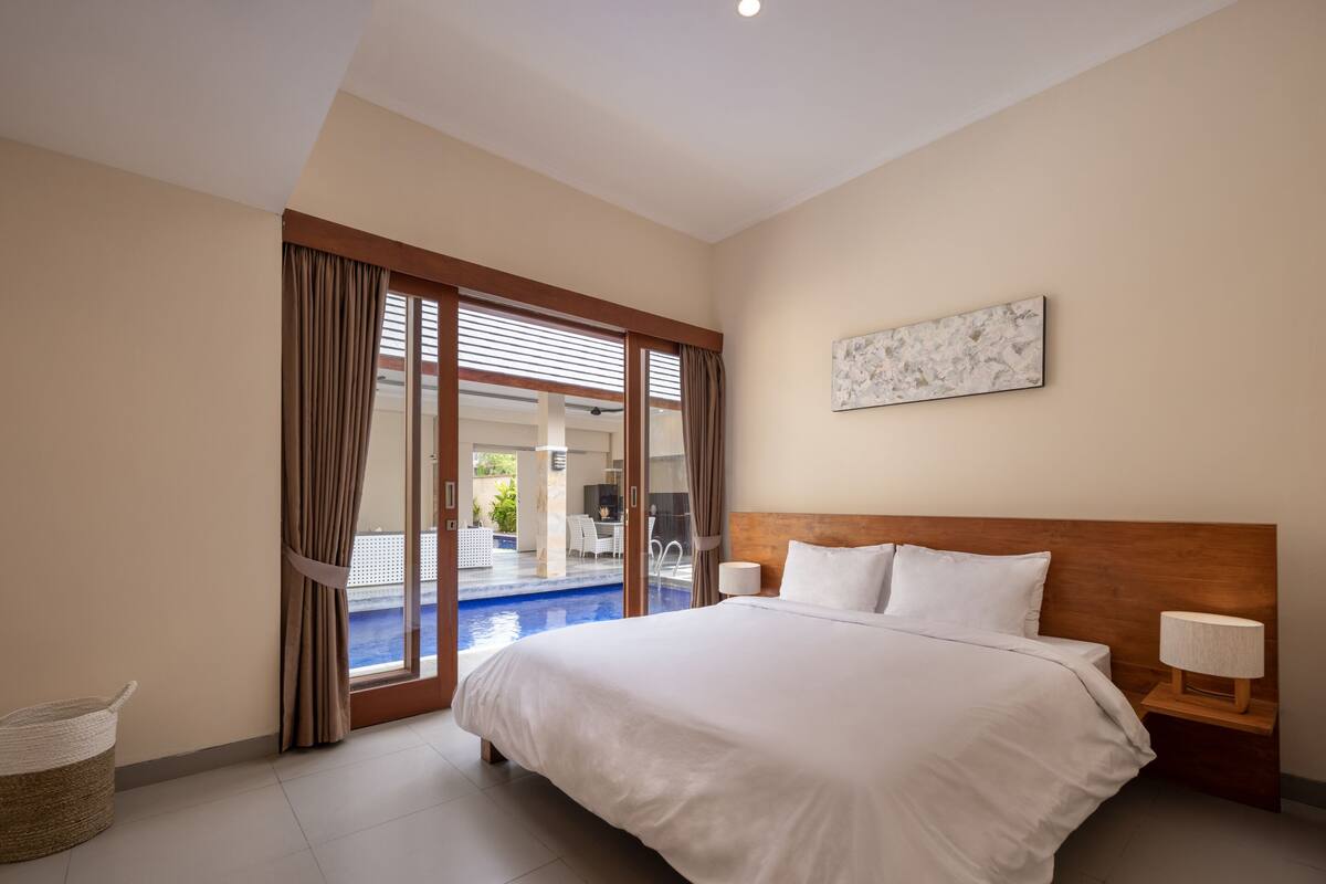 Quarto de hotel com uma cama grande, cabeceira de madeira, um quadro na parede e vista para a área da piscina.