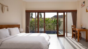 Une chambre avec un grand lit, une table de chevet en bois, un bureau et une vue sur un jardin, accessible par une porte-fenêtre coulissante.