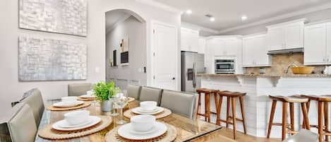A modern kitchen with white cabinets, a marble countertop, and a dining area with a glass table set for six.