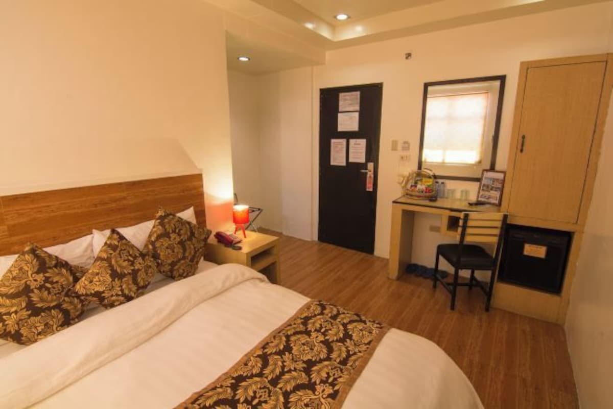Una habitación de hotel con cama, mesita de noche con lámpara, escritorio con silla, armario y puerta con documentos.