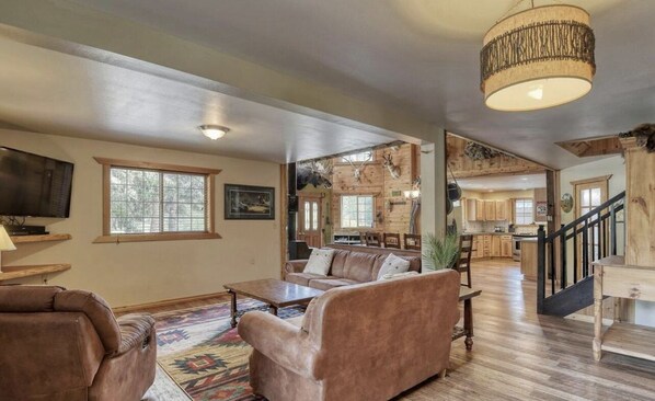 Un salón espacioso con un sofá grande, una mesa de centro y un televisor. Se ve una escalera que conduce a una zona de cocina con armarios de madera y un candelabro.