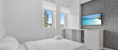 A modern bedroom with a bed, a TV mounted on a grey wall, and a window with a view of a beach.