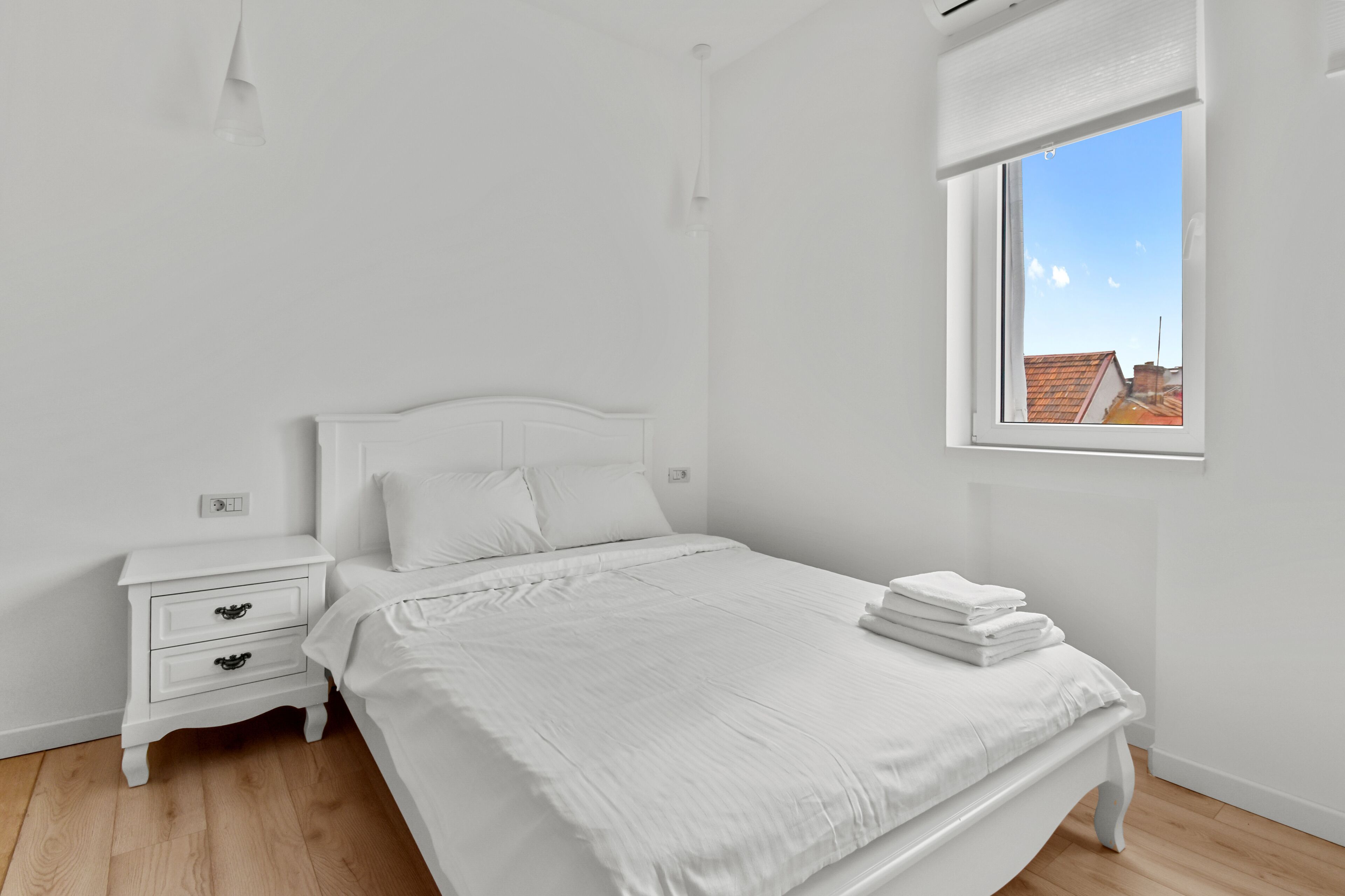 A white bedroom with a bed, a nightstand, a window with a view of buildings, and a ceiling light.