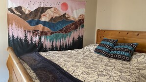 A bed with a patterned bedspread, two patterned pillows, a dark throw blanket, and a tapestry depicting a mountainous landscape with birds.