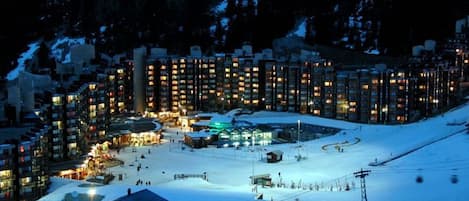 A snow-covered ski resort at night with illuminated buildings and a ski lift.