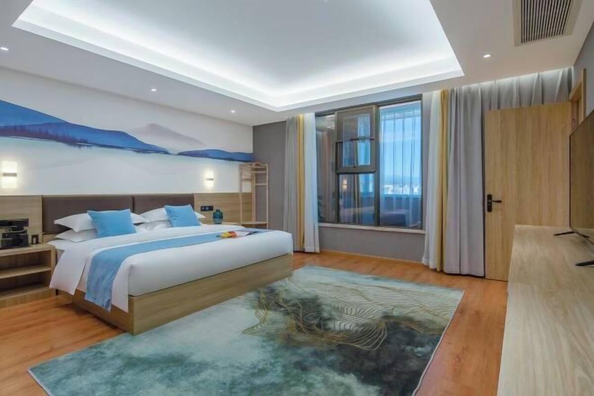 A modern hotel room with a large bed, a desk, a window with curtains, and a ceiling light.