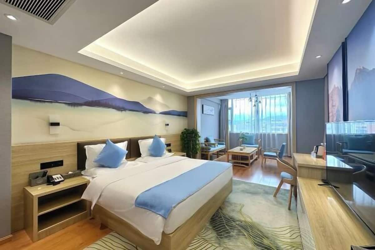 A modern hotel room with a large bed, bedside tables, a desk, a chair, a TV, and a view of the city through large windows.