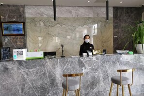 A reception desk with a marble counter, a framed menu, and a digital screen. There are two chairs with wooden legs and cushioned seats.