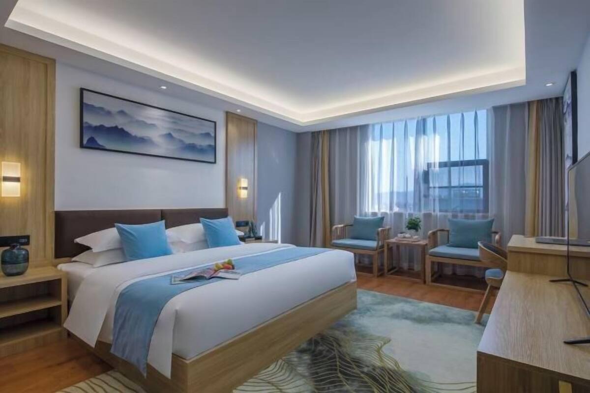 A modern hotel room with a large bed, a desk, and a chair. There is a painting on the wall and a window with curtains.