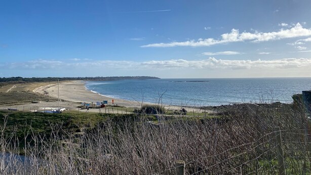 Una spiaggia costiera con cielo sereno, una battigia sabbiosa e alcune strutture in lontananza.