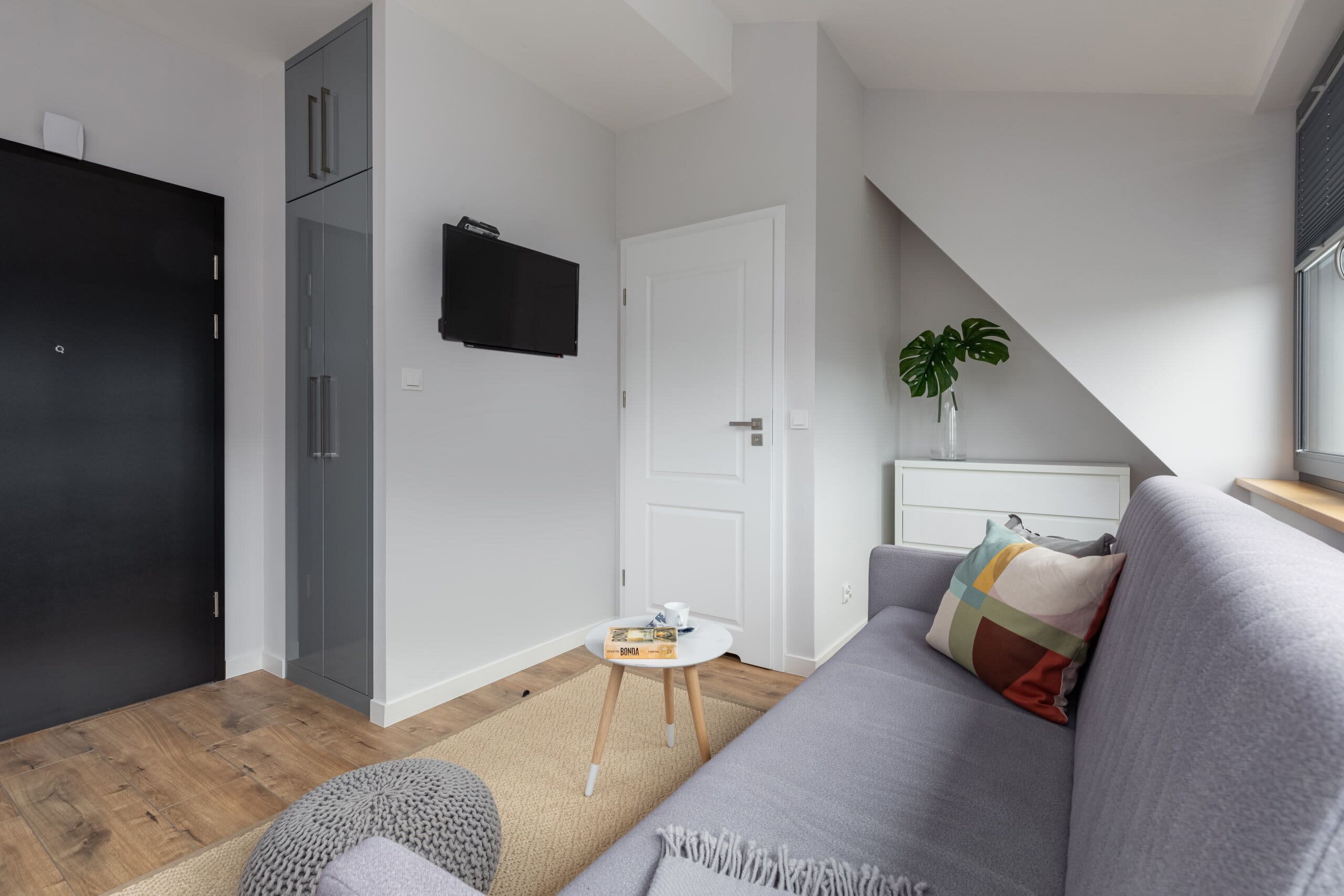 Una sala moderna con un sofá gris, una mesita redonda con un libro y una taza, un mueble negro y un televisor en la pared.