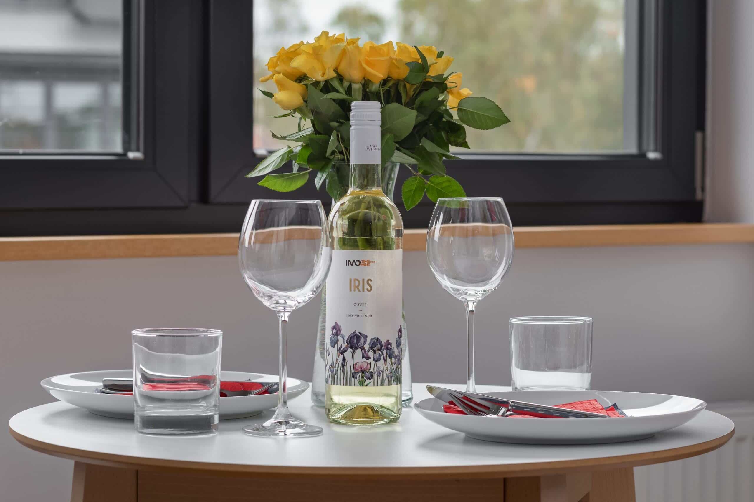 A table set with wine glasses, a bottle of Iris wine, and a vase of yellow roses.