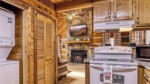 A cozy cabin-style kitchen with wooden walls, a stone fireplace, a television mounted above it, and a ceiling fan. - Lacey's Place by Beyond Expectations: Dog-Friendly Cabin w/ Hot Tub & Fire Pit (Sevierville)