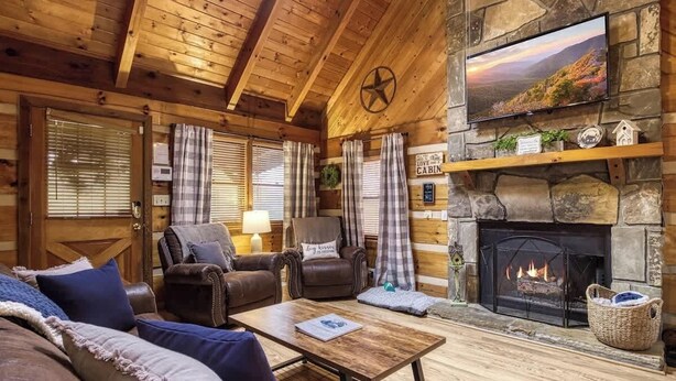 A cozy cabin interior with a stone fireplace, wooden walls, and a television mounted above it. - Lacey's Place by Beyond Expectations: Dog-Friendly Cabin w/ Hot Tub & Fire Pit (Sevierville)