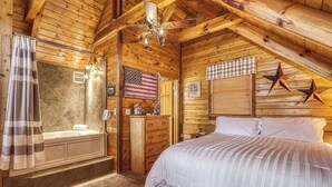 A cozy log cabin bedroom with a large bed, a wooden dresser, a vanity, and a shower area. - Lacey's Place by Beyond Expectations: Dog-Friendly Cabin w/ Hot Tub & Fire Pit (Sevierville)