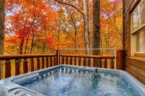 Un jacuzzi con cubierta de madera y barandilla, rodeado de árboles con follaje otoñal.