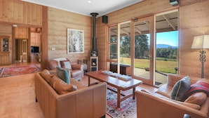 A spacious living room with a fireplace, leather sofas, a wooden coffee table, and large windows offering a view of the outdoors.