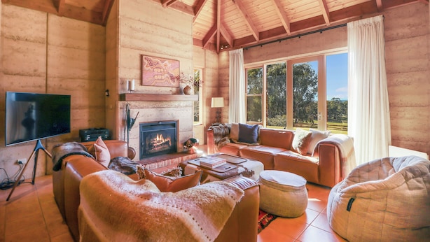 A cozy log cabin interior with a fireplace, leather sofas, a bean bag, and a large window offering a view of the outdoors.