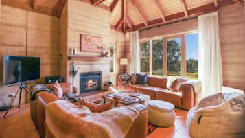 A cozy log cabin interior with a fireplace, leather sofas, a bean bag, and a large window offering a view of the outdoors.