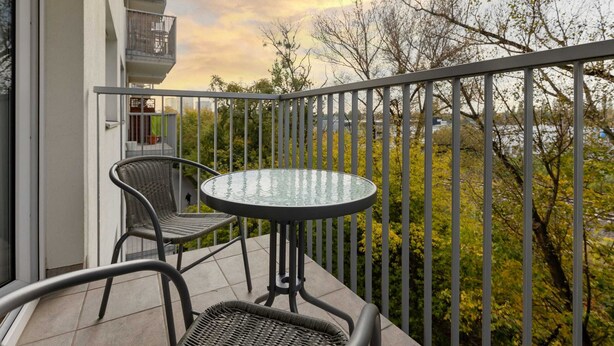 Un balcon doté d’une table ronde en verre et de deux fauteuils en osier, donnant sur une zone bordée d’arbres.