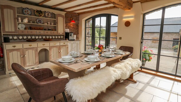 A dining area with a wooden table set for four, surrounded by cushioned chairs. A stone wall and a large window provide natural light. - Pen Bont Home Farm (Brecon)