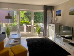 Un salon moderne avec un téléviseur à écran plat, un canapé, une table basse et un tapis noir. Une porte-fenêtre coulissante donne accès à un balcon agrémenté de plantes et de mobilier d’extérieur.