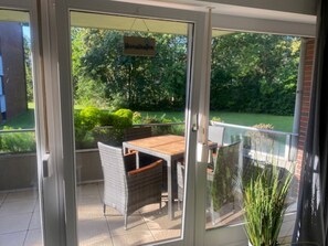 En glasdør fører ud til en udendørs terrasse med borde og stole, et hængende skilt og udsigt til grønne områder.