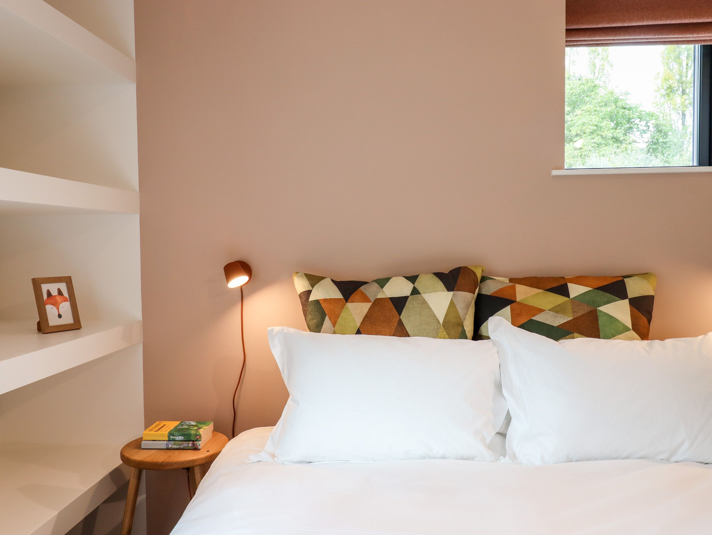 Een slaapkamer met een bed, kussens, een nachtkastje, een lamp, een ingelijst beeld en een raam met uitzicht op bomen.
