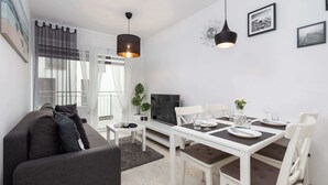 A modern living room with a sofa, a dining table set for four, and a TV. - Modern Apartment | Separate Bedroom and Balcony (Kołobrzeg)