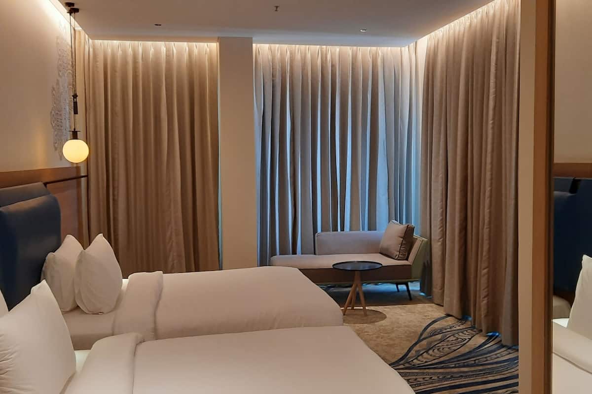 Quarto de hotel com uma cama grande, um sofá, uma mesa redonda pequena e vista através de cortinas leves.