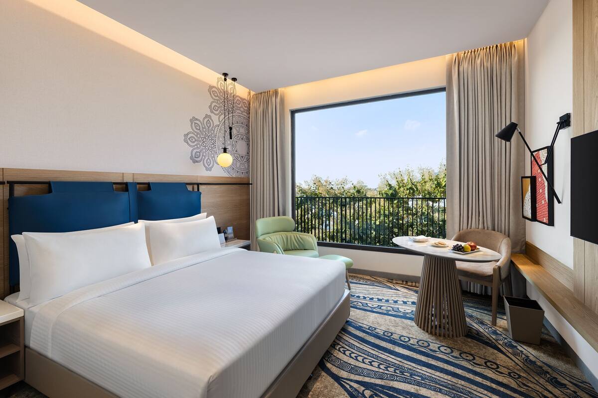Quarto de hotel com uma cama grande, uma mesa pequena, uma cadeira, televisão e varanda com vista.