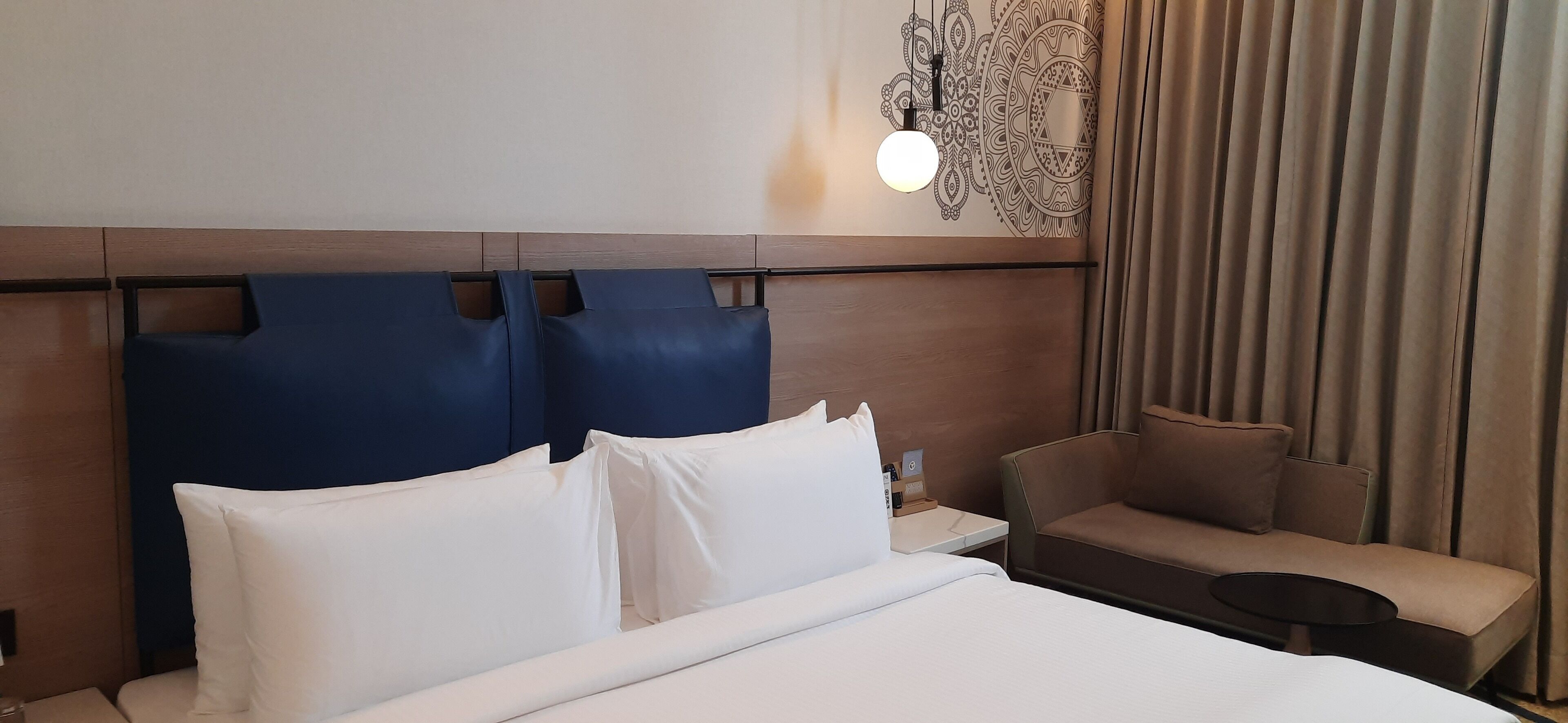 A hotel room with a large bed, a blue headboard, a small sofa, a nightstand, and a wall-mounted light fixture.