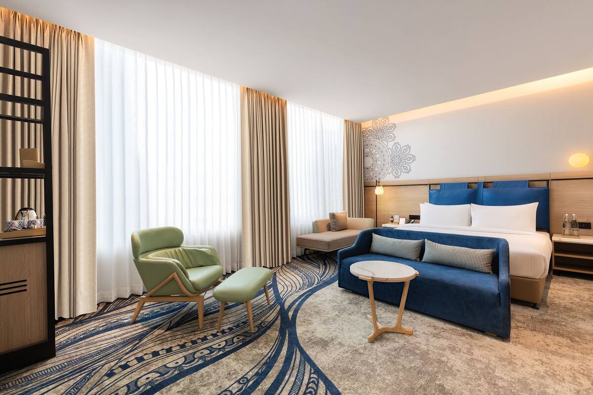 Quarto de hotel com uma cama, um sofá, uma poltrona verde, uma mesinha e um tapete estampado.