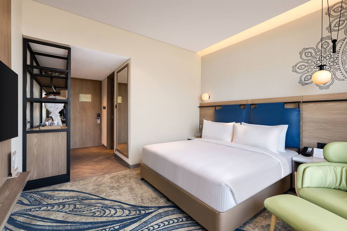 Quarto de hotel com uma cama grande, uma cadeira verde, uma escrivaninha com abajur e um guarda-roupa.